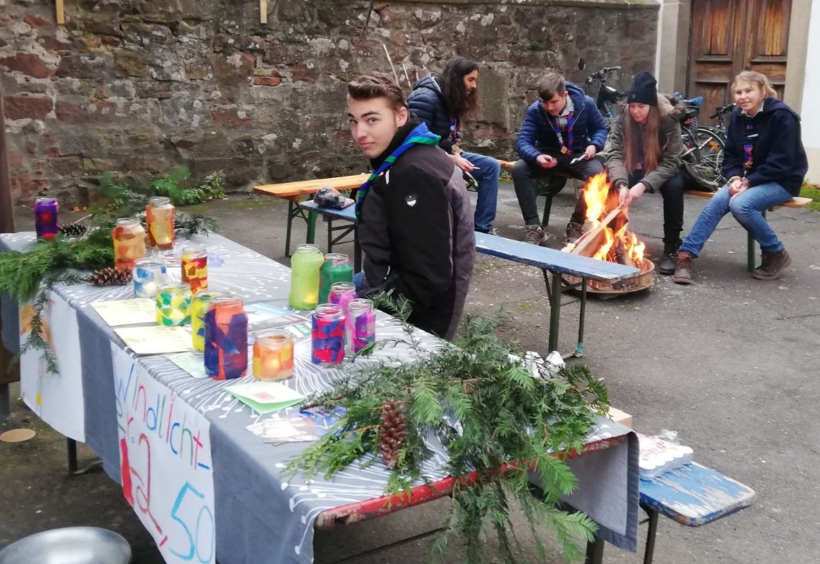 Verkaufstisch mit Windlichtern und Tannenzweigen. Im Hintergrund Leute um ein Lagerfeuer